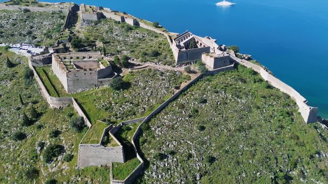 Scenic 4K drone panning shot right of Palamidi Fortress in Nafplio, Greece. Revealing massive stone castle walls on a high ridge overlooking a calm blue sea with a luxury yacht.