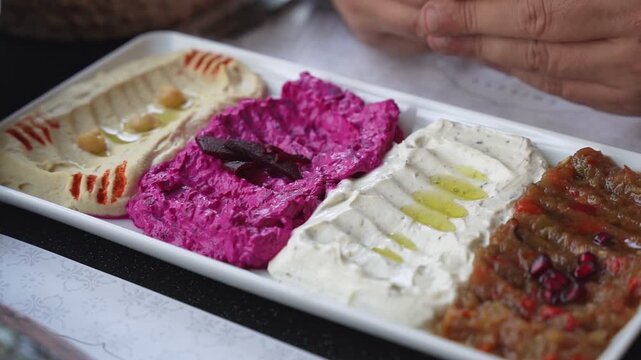 Close-up of a colorful Mediterranean mezze platter featuring hummus, beetroot dip, and baba ganoush on a restaurant table. A person waits to eat this healthy Middle Eastern appetizer selection.