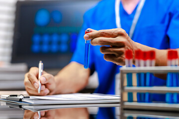 Medical lab technician in blue scrubs holding test tube and writing notes beside rack of blood samples in laboratory, healthcare diagnostics research concept, shallow depth of field. © M+Isolation+Photo