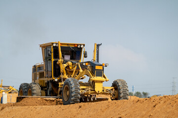 Fototapeta premium Road Grader on Earth: A heavy-duty yellow road grader with large tires levels a mound of dirt at a construction site under a clear sky, showcasing industrial progress.