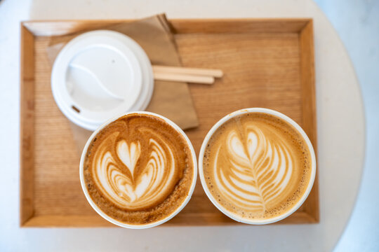 A wooden tray holds two beautifully crafted lattes with intricate heart designs, placed next to a white ceramic cup with a lid. The scene captures the artistry of coffee making, emphasizing the