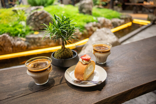 A wooden table hosts two glasses of iced coffee, a plate with a sandwich, and a small potted plant. The scene is set in a serene garden with a stone path and lush greenery. The warm lighting and