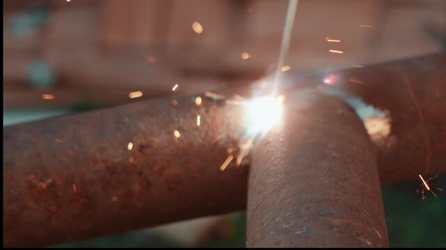 Close up of professional welder joining steel pipes with electric arc outdoors. Flying orange sparks and dramatic light create powerful heavy industry and engineering concept. Poor quality welding