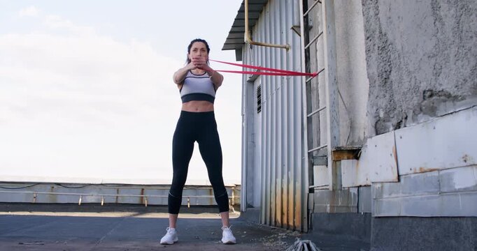 Athletic motivation. Stretching band workout. Slim female trainer showing intensive effort training extending red elastic loop on rooftop outside.