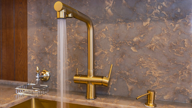 A gold-colored metal faucet on the edge of the sink. The water flows in thin streams of the shower. A soap dish, a dispenser. The countertop, wall panel are made of artificial stone quartz agglomerate