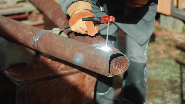 Closeup of professional welder joining steel pipes in outdoor workshop area. Protective gear, glowing sparks and industrial atmosphere highlight technical skills and durable metal construction.