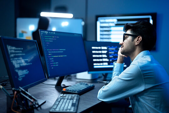A Stressed programmer has headache while writing program code on multiple desktop screens while his colleague working in the background in a modern office.