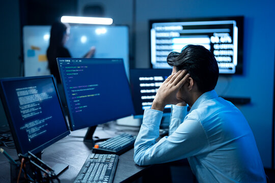 A Stressed programmer has headache while writing program code on multiple desktop screens while his colleague working in the background in a modern office.
