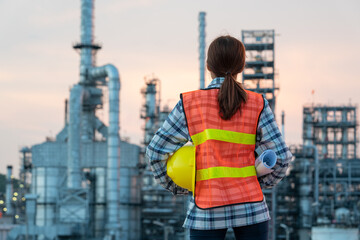 Industrial Powerhouse: A female engineer stands before a sprawling petrochemical plant, holding a hard hat and blueprints, symbolizing innovation and dedication in the industrial sector. © Rassarin