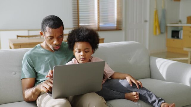 African father and son learning together with laptop on sofa at home.