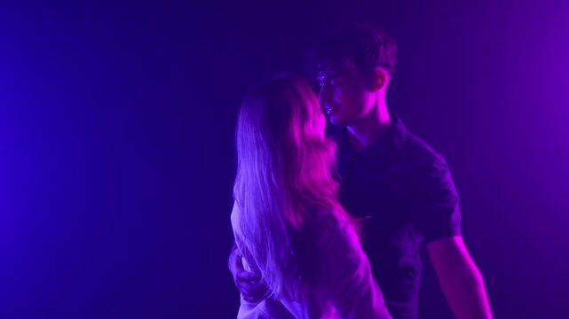 Romantic couple dancing in neon light, embodying love and intimacy. A young man and woman embrace in a dark club setting, illuminated by vibrant purple and blue hues. This footage is projects related 