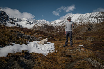 Man hiker with wooden stick and white dog resting on snow in mountains. Male traveler with husky looking at pet, trekking in alpine valley, outdoor travel adventure and lifestyle concept © ANDREY