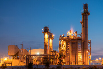 Power Generation Facility at Dusk: A large industrial power plant with towering smokestacks and illuminated machinery stands against the twilight sky. © Rassarin