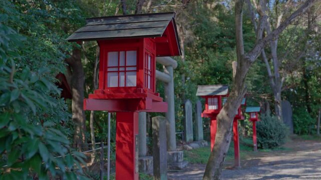 A close perspective of a vivid red wooden shrine lantern leads the eye down a row of identical lanterns receding along a shaded forest path, with stone pillars and dense green woodland creating a rhyt