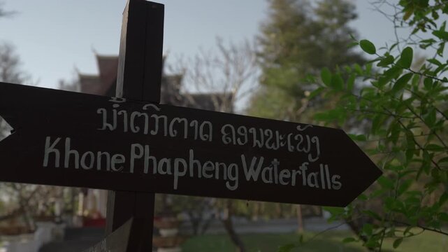 Close-up slow moving shot of a wooden Khone Phapheng Waterfalls direction sign, with foliage and a blurred temple structure in the background.