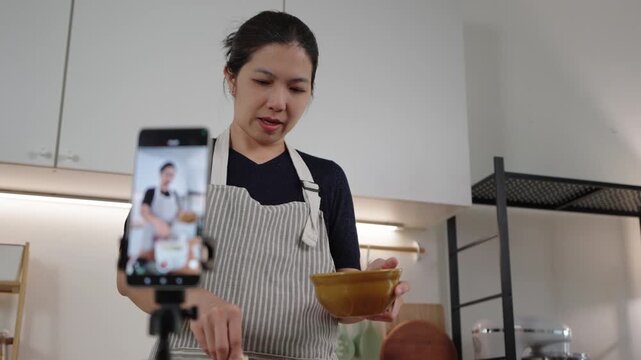 Asian woman recording cooking video with smartphone in modern kitchen.