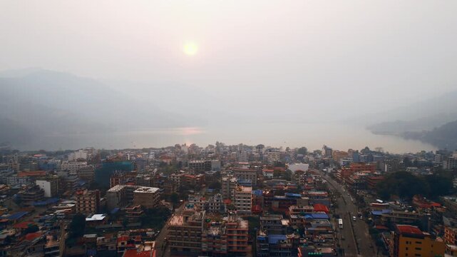 An aerial drone view captures the dense urban sprawl of Pokhara city stretching along the misty shores of Fewa Lake under a hazy Nepalese sky.