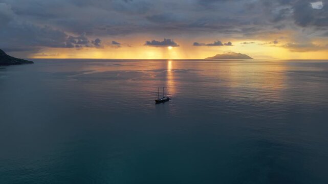 Cinematic drone footage flying towards a sailing boat on calm waters at sunset. Features dramatic golden clouds and sun rays over the Indian Ocean in Beau Vallon, Mahe. Seychelles 30fps 001