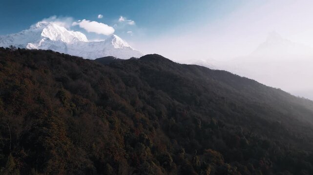 An aerial view over a forested ridge reveals the snow-capped Annapurna range and Fishtail peak rising above drifting clouds in Nepal.
