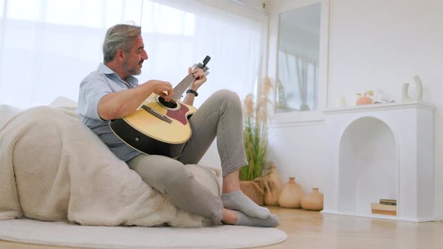 Mature man relaxing at home while playing acoustic guitar on sofa.