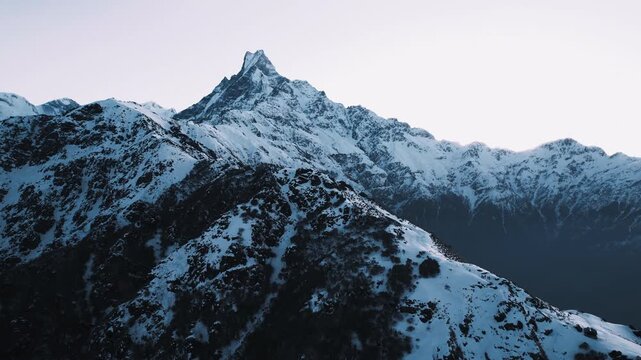 Aerial drone view of the sharp snow-capped Machapuchare peak rising above a rugged Himalayan ridge at twilight in Nepal.