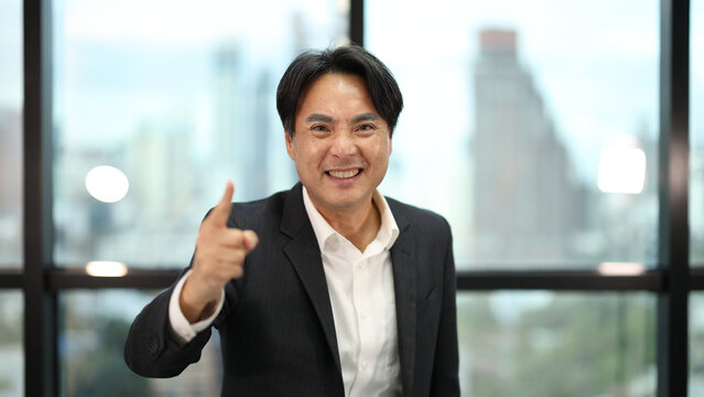 Smiling Asian Businessman Pointing Finger at Camera in Modern Office Setting