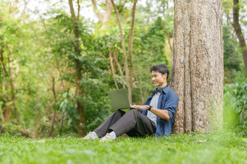 Obraz premium Asian man working remotely on laptop while sitting against tree in outdoor park