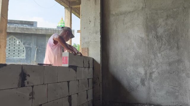 Instructional-quality footage of a construction worker carefully placing a hebel (AAC) block onto a pre-laid layer of wet cement mortar on top of an existing wall