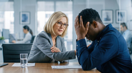 Fototapeta premium A blonde woman in a grey blazer comforts a stressed man holding his head at a conference table in a modern office while colleagues work in the blurred background