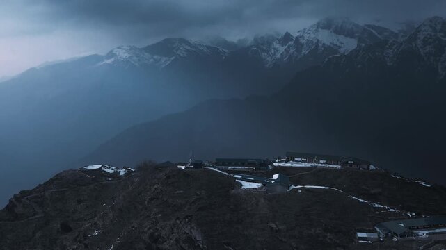 Atmospheric drone shot of a remote mountain lodge complex perched on a high Himalayan ridge in Nepal, with dramatic moody clouds rolling across snow-dusted peaks.