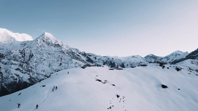 Stunning aerial view of trekkers walking along a snow-covered Himalayan ridge in Nepal, with the massive Annapurna peaks towering against a clear blue sky.
