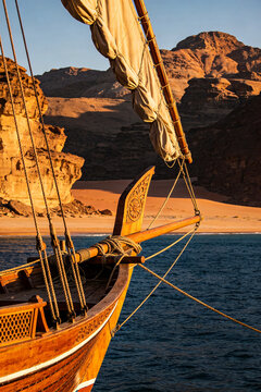 Traditional wooden dhow boat sailing near rugged desert mountains during a beautiful golden sunset hour