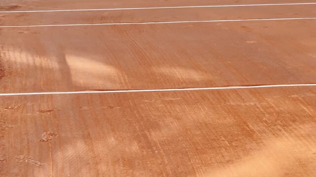 Close-up of an empty clay tennis court beside the net, with white boundary lines, groomed orange surface, and subtle footprints in warm natural light.