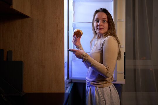Female holding muffin near refrigerator turning to camera indoors