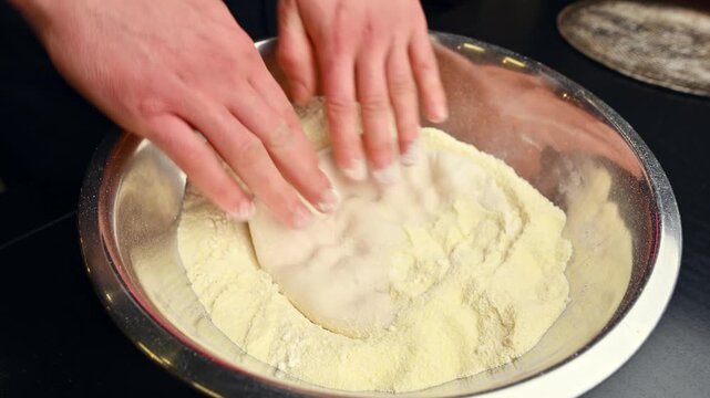 Hands knead dough in bowl with flour, preparing for baking process in kitchen