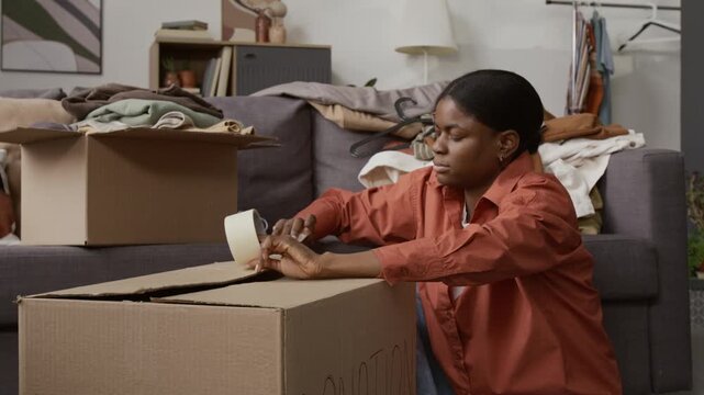 Tilt down view of Black gen Z girl sealing cardboard box with adhesive tape while packing stuff for donation
