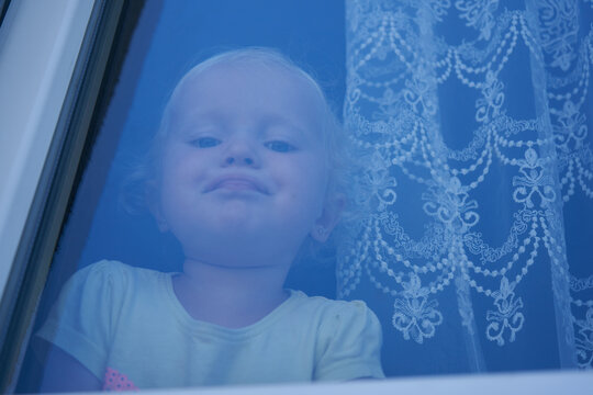 Cute little blonde toddler girl looking through a window glass, Portrait of a blond girl sad or waiting by a window with a white tulle curtain.