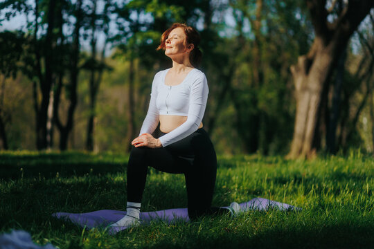 Mature woman doing fitness meditating with closed eyes in sunny forest park