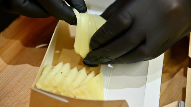 Hands in black gloves arranging sliced cheese in a cardboard container on wooden surface