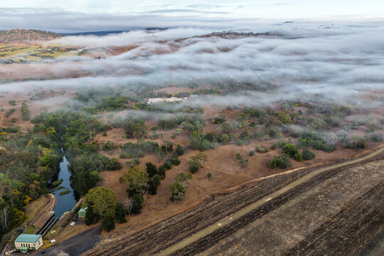 Aerial image of the Glenbawn Dam wall with morning fog drifting through the surrounding valley in NSW Australia. Drone photograph captures the reservoir, rugged mountain landscape