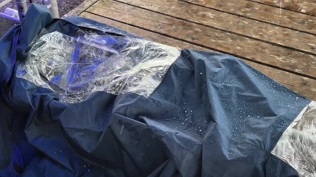 Close-up of a waterproof protective tarp covering outdoor furniture during hailstorm, showing raindrops and weather protection concept on a wooden terrace.