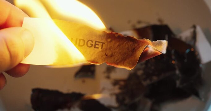 A close-up slow-motion shot of a piece of paper labeled &ldquo;BUDGET&rdquo; burning. The flame consumes the paper gradually, symbolizing financial loss, overspending, or waste.