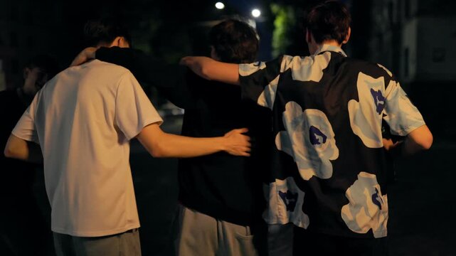 Four teenage boys walking arm in arm down a city street at night from behind.