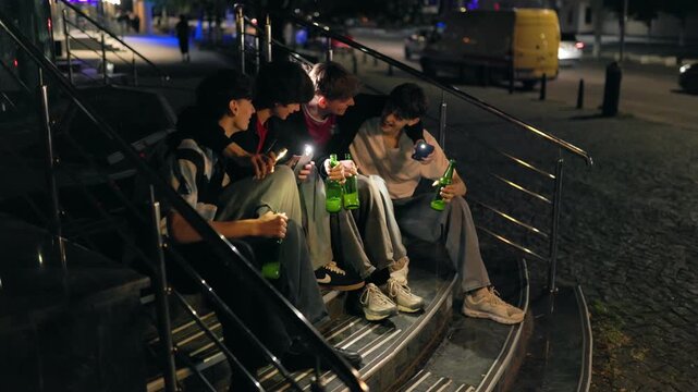 Four happy teenage boys using phone screen flashlights on stairs at night.