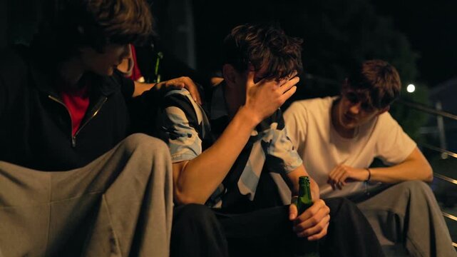 Close up of a depressed teenage boy crying while friends comfort him at night.