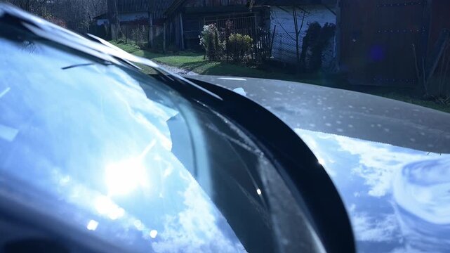Wipers wipe the windshield of an SUV. Sunlight sparkles like diamonds on a car's windshield, capturing reflections of the vibrant sky and scenic landscape in a peaceful village setting.