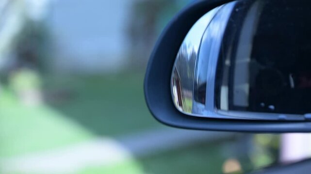 Rear view mirror adjustment. A car's side mirror captures reflections of a peaceful suburban scene, highlighting vibrant greenery and blue skies on a calm afternoon drive.