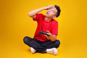 Exhausted Young Asian Man Sitting Cross legged Holding Gamepad and wiping Sweat from His Forehead © Gatot