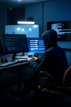 Male computer hacker in black hoodie typing code on computer in a dark room with light reflection on glasses.