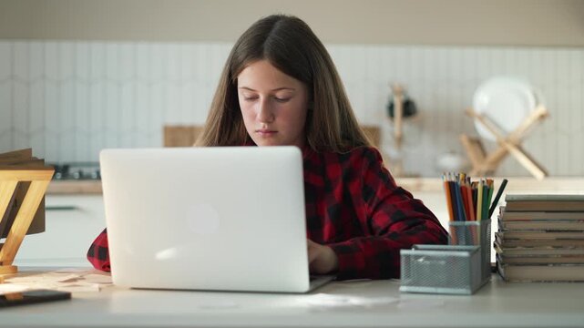 Teenage girl studies at home using laptop computer. Student types on keyboard while doing homework. Young schoolgirl focuses on online learning class. Books and pencils rest on desk near laptop.